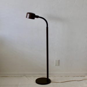 以下が含まれることがあります： カーブした首と円形のベースを備えた、濃い茶色のフロアランプ。 ランプは円筒形のシェードと長くまっすぐなポールを備えています。 ランプはコンセントに差し込まれています。