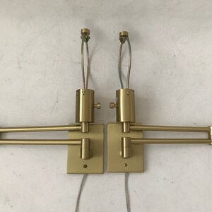 May include: A pair of gold-toned wall-mounted swing arm lamps with adjustable arms. The lamps have a simple, modern design and are perfect for reading or task lighting.