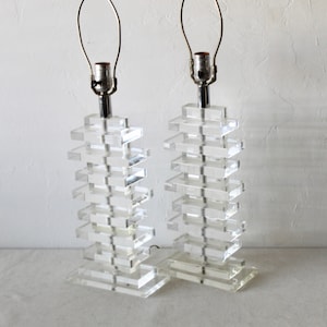 May include: A pair of clear acrylic table lamps with a stacked design. Each lamp has a silver metal base and a clear glass shade.