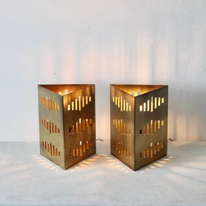 Puede incluir: Dos lámparas geométricas doradas, con forma de prisma triangular. Cada lámpara tiene una serie de cortes verticales que permiten que la luz brille. Las lámparas están encendidas, proyectando un brillo cálido y creando un efecto de iluminación decorativo.