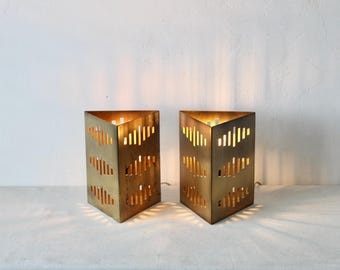 Pode incluir: Duas lâmpadas geométricas douradas, com formato de prisma triangular. Cada lâmpada possui uma série de recortes verticais que permitem que a luz brilhe. As lâmpadas estão acesas, projetando um brilho quente e criando um efeito de iluminação decorativo.