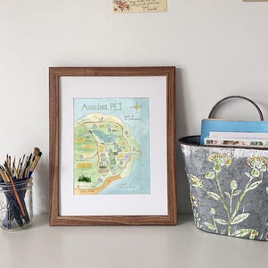 May include: A framed watercolour map of Avonlea, PEI, in a brown wooden frame. The map shows a detailed illustration of the area, including landmarks and the coastline. A mason jar with paintbrushes and a decorative metal bucket are also visible.