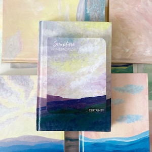 May include: A small notebook with the text "Scripture to Memorize" and the word "Certainty" on the cover. The cover features a landscape painting with blue, purple, and green hues. The notebook is surrounded by other painted artworks.
