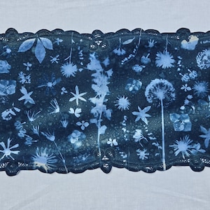 May include: A rectangular fabric runner featuring a dark blue cyanotype print of various botanical silhouettes, including dandelion seed heads, leaves, and small flowers, finished with a decorative dark blue scalloped lace border.