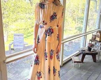 May include: A cream-colored, vintage-style dress with a floral pattern in red, blue, and purple. The dress features short sleeves, a collared neckline, and a matching belt. The dress is floor-length and worn with black boots.