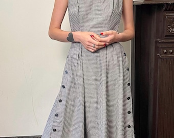 May include: A sleeveless midi dress in a grey and white micro-check pattern featuring a square neckline and a row of dark buttons running down each side of the skirt. The dress has a fitted bodice and a flared skirt that falls below the knees.