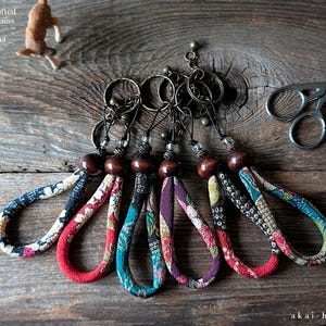May include: Japanese handcrafted Chirimen keychains with colorful fabric loops and wooden beads. The keychains are arranged on a rustic wooden surface, with a small deer figurine and decorative scissors nearby. The text "akai-hana.com" is visible.