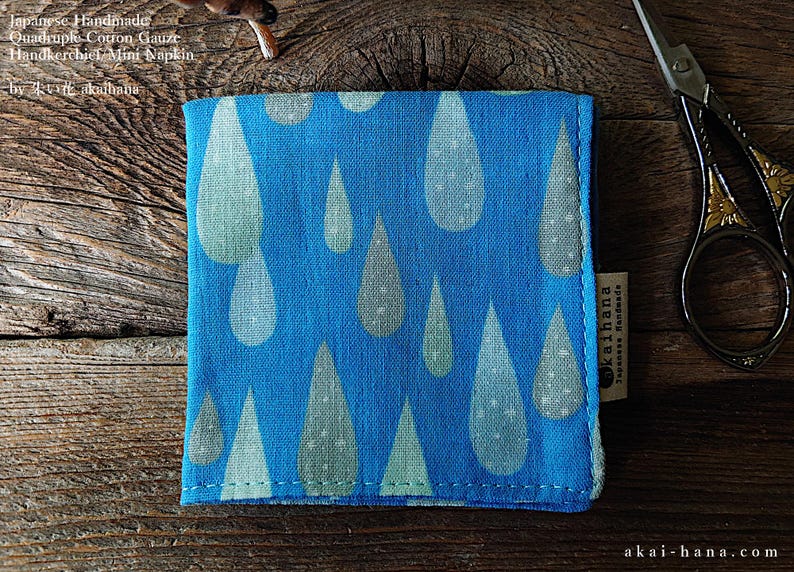 May include: A square blue cotton gauze handkerchief featuring a repeating pattern of light green and white raindrops. The fabric has a visible woven texture and a small side tag labeled akaihana. The text reads Japanese Handmade Quadruple-Cotton Gauze Handkerchief Mini Napkin by akaihana.