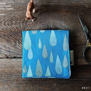 May include: A square blue cotton gauze handkerchief featuring a repeating pattern of light blue and white raindrops. The fabric has a small side tag with text and is folded neatly. The text on the image reads: Japanese Handmade Quadruple Cotton Gauze Handkerchief/Mini Napkin by akaihana.