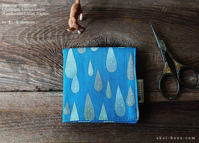 May include: A square blue cotton gauze handkerchief featuring a repeating pattern of light blue and white raindrops. The fabric has a small side tag with text and is folded neatly. The text on the image reads: Japanese Handmade Quadruple Cotton Gauze Handkerchief/Mini Napkin by akaihana.