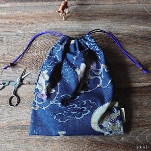 以下が含まれることがあります： 紺色の巾着袋。装飾的な模様と紫色の紐が付いています。袋は布製で、側面にタグが付いています。「Japanese Handmade KINCHAKU Drawstring Purse」の文字が見えます。ハサミと小さな置物も写っています。