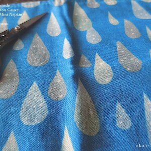 May include: A blue cotton gauze handkerchief featuring a repeating pattern of light-colored raindrops. The fabric is labeled Japanese Handmade Quadruple Cotton Gauze Handkerchief/Mini Napkin by akaihana. A pair of black metal scissors rests on the fabric.