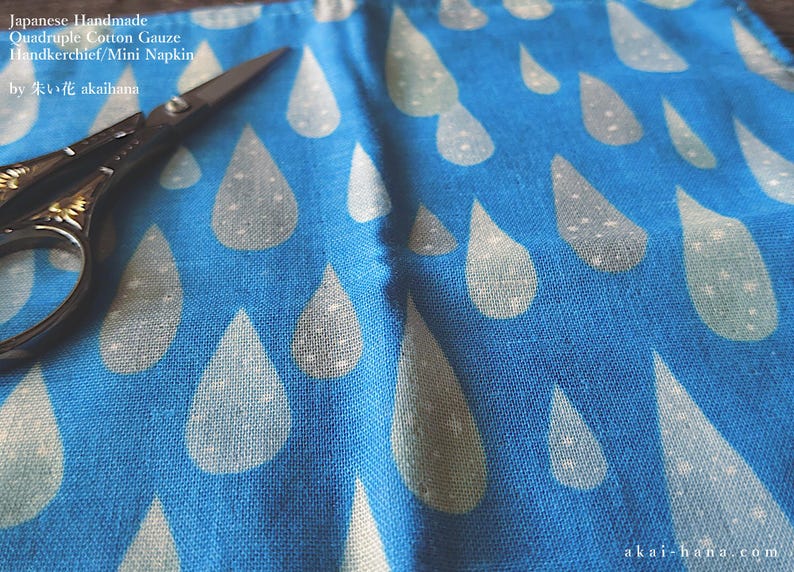 May include: A blue cotton gauze handkerchief featuring a repeating pattern of light-colored raindrops. The fabric is labeled Japanese Handmade Quadruple Cotton Gauze Handkerchief/Mini Napkin by akaihana. A pair of black metal scissors rests on the fabric.