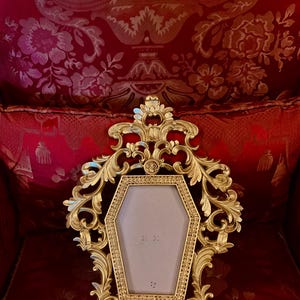 May include: An ornate gold-coloured picture frame shaped like a coffin. The frame has a detailed design with scrolls and flourishes. The frame is sitting on a red velvet surface.
