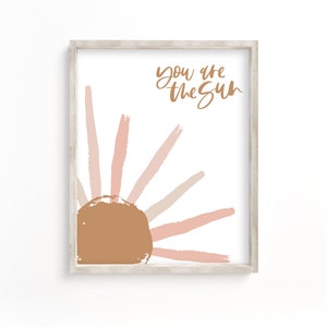 Può includere: Una stampa incorniciata con un design minimalista del sole in tonalità rosa e marrone. Il testo "You are the sun" è scritto in corsivo.