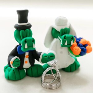 May include: A whimsical wedding cake topper featuring two green alligator figures. One alligator wears a black tuxedo and top hat, the other a white wedding dress and holds a bouquet. A silver wedding ring set is in the foreground.