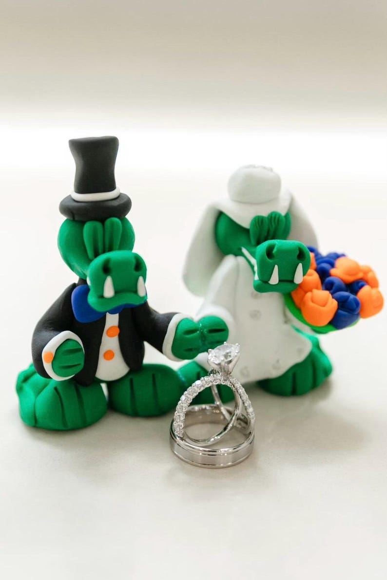 May include: A whimsical wedding cake topper featuring two green alligator figures. One alligator wears a black tuxedo and top hat, the other a white wedding dress and holds a bouquet. A silver wedding ring set is in the foreground.