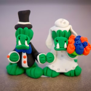 May include: Clay figurines of a bride and groom, both with green reptilian faces. The groom wears a black tuxedo and top hat. The bride holds a bouquet of orange, blue, and green flowers and wears a white dress and veil. Wedding theme.