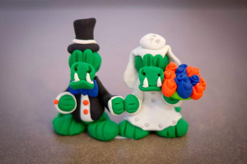 May include: Clay figurines of a bride and groom, both with green reptilian faces. The groom wears a black tuxedo and top hat. The bride holds a bouquet of orange, blue, and green flowers and wears a white dress and veil. Wedding theme.
