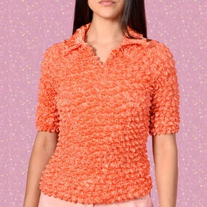 Può includere: Una camicia a maniche corte di colore corallo con un tessuto strutturato e stropicciato. La camicia ha un colletto e una chiusura con bottoni.