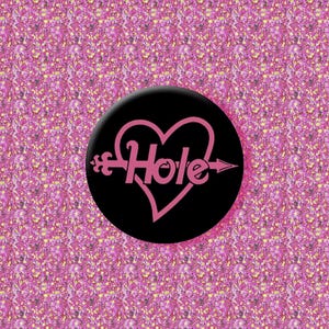 Puede incluir: Un botón negro con la palabra "Hole" en rosa, con un diseño de corazón y flecha rosa. El botón está sobre un fondo floral rosa.