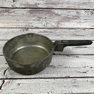 Op de afbeelding: Een vintage metalen steelpan met een lange, zwarte handgreep. De pan heeft een doffe zilverkleur en lijkt goed gebruikt. De handgreep is gemaakt van een donker, mogelijk plastic materiaal. De pan staat op een verweerd, wit geschilderd houten oppervlak.
