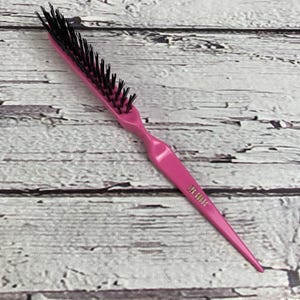 May include: A pink hairbrush with black bristles. The brush has a long, pointed handle and a curved design for easy grip. The text "SYR GROOM" is printed on the handle. The brush is designed for detangling and styling hair.