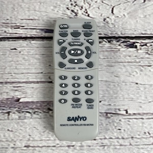 Peut inclure: Une télécommande Sanyo grise avec de nombreux boutons pour diverses fonctions, notamment l'alimentation, le volume et le réglage. La télécommande est rectangulaire et porte la marque "SANYO" en bas.