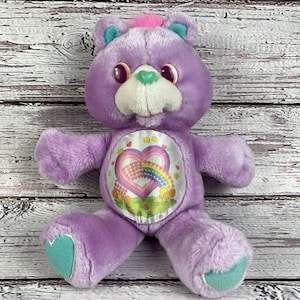 May include: A vintage Care Bear plush toy in lavender with a heart-shaped emblem on its belly. The bear has a white muzzle, a green heart-shaped nose, and blue accents on its ears and feet. A pink tuft of hair sits atop its head.