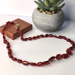 Puede incluir: Un collar de ámbar rojo con un cierre de oro. El collar está hecho de cuentas ovaladas y está sobre una superficie marrón.