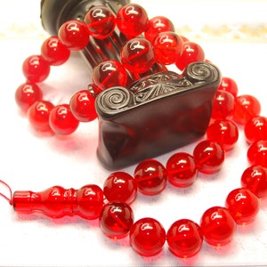 Könnte beinhalten: Eine rote Glas-Gebetskette mit einem Quastenanhänger. Die Perlen sind rund und haben eine glänzende Oberfläche. Die Kette liegt auf einer weißen Oberfläche.