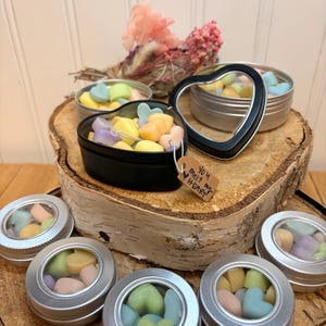 May include: A collection of heart-shaped wax melts in pastel colors, displayed in small, round tins with clear lids and a black heart-shaped tin. The melts are arranged on a wooden slice, with a tag that reads "You melt my heart."