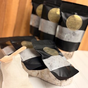 Puede incluir: Se muestran varias bolsas negras con una franja plateada y una etiqueta dorada. Una peque&ntilde;a cuchara de madera descansa sobre una tela blanca. Las bolsas contienen una sustancia granular.