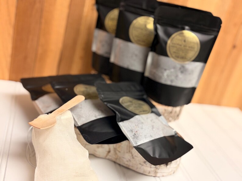Puede incluir: Se muestran varias bolsas negras con una franja plateada y una etiqueta dorada. Una peque&ntilde;a cuchara de madera descansa sobre una tela blanca. Las bolsas contienen una sustancia granular.