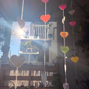 May include: A string of colorful heart-shaped decorations hanging in front of a porch. The hearts are various colors, including orange, pink, and yellow, and are suspended from a white string. The background includes a wooden pallet and plants.