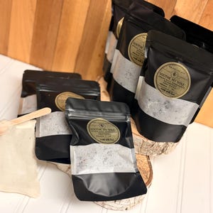 Puede incluir: Bolsas negras con una etiqueta dorada que dice "Soothe My Sole" que contienen sales de ba&ntilde;o. Tambi&eacute;n se incluye una peque&ntilde;a cuchara de madera y una peque&ntilde;a bolsa de tela. Las bolsas se exhiben sobre una superficie de madera.