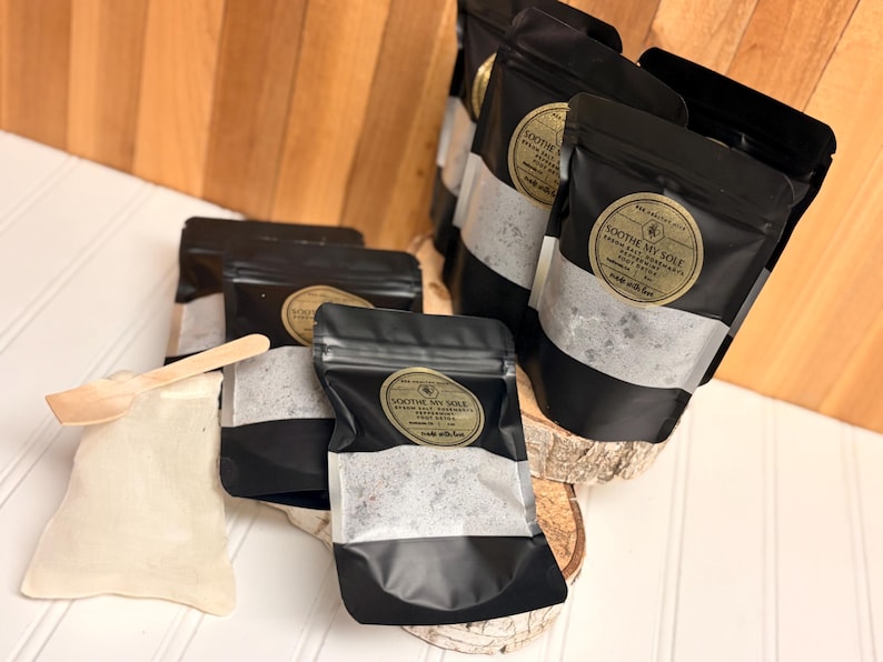 Puede incluir: Bolsas negras con una etiqueta dorada que dice "Soothe My Sole" que contienen sales de ba&ntilde;o. Tambi&eacute;n se incluye una peque&ntilde;a cuchara de madera y una peque&ntilde;a bolsa de tela. Las bolsas se exhiben sobre una superficie de madera.