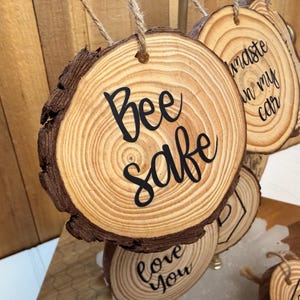 May include: Wooden slice ornaments with natural bark edges. The front of the ornaments have black text that reads "Bee safe", "Waste in my car", and "Love you". Each ornament is suspended by a natural twine.