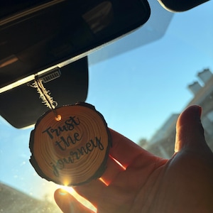 以下が含まれることがあります： 「Trust the journey」の文字が黒い筆記体で書かれた、丸い木の車の芳香剤。芳香剤は木の枝の断面から作られており、木の自然な木目が見えます。紐で吊り下げられており、車のフロントガラスの前に掲げられています。