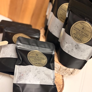 Puede incluir: Varios sobres negros con etiquetas doradas y forro de papel blanco, llenos de una sustancia granular. Las etiquetas dicen "Soothe My Sole" con detalles del producto. Los sobres est&aacute;n dispuestos sobre una superficie de madera y sellados.