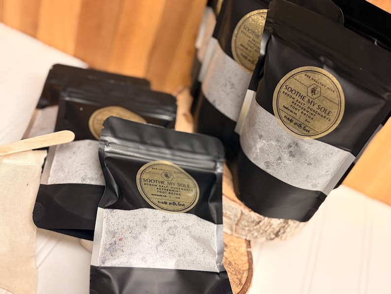 Puede incluir: Varios sobres negros con etiquetas doradas y forro de papel blanco, llenos de una sustancia granular. Las etiquetas dicen "Soothe My Sole" con detalles del producto. Los sobres est&aacute;n dispuestos sobre una superficie de madera y sellados.