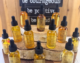 May include: A collection of eleven small amber glass dropper bottles containing various oils, each with a black dropper cap and a label. The bottles are arranged on tiered wooden platforms, with a sign in the back reading be courageous be positive.