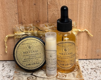 May include: A collection of Bee Healthy Hive products, including a round tin of beeswax salve, a lip balm, and a dropper bottle of tincture. The items are displayed on a decorative gold-colored fabric and set against a wooden background.