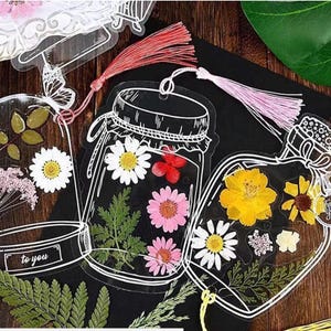 May include: Clear acrylic jar-shaped bookmarks with pressed flowers and decorative tassels. The bookmarks feature white outlines and include daisies, pink flowers, and yellow flowers. One bookmark has the text "to you".