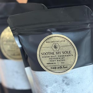 Puede incluir: Bolsas negras con una etiqueta dorada que dice "SOOTHE MY SOLE" con sal de Epsom, romero y menta. La etiqueta tambi&eacute;n dice "BEE HEALTHY HIVE" y "FOOT DETOX". Las bolsas est&aacute;n llenas de un polvo blanco.