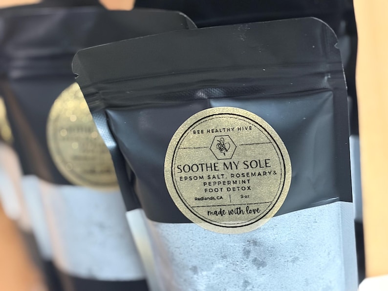 Puede incluir: Bolsas negras con una etiqueta dorada que dice "SOOTHE MY SOLE" con sal de Epsom, romero y menta. La etiqueta tambi&eacute;n dice "BEE HEALTHY HIVE" y "FOOT DETOX". Las bolsas est&aacute;n llenas de un polvo blanco.
