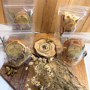 May include: An assortment of dried herbs and botanicals in clear pouches, arranged on a wooden surface. The pouches are labeled with text such as "Still & Sacred," "Harvest Hearth," "Bloom & Breathe," and "Golden Hour." A wooden bee and a wooden slice with "bee healthy hive creations" are also present.