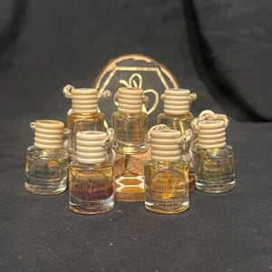 Könnte beinhalten: Eine Sammlung von acht kleinen Glasflaschen mit Korkstopfen und Etiketten. Die Flaschen enthalten natürliche Auto-Lufterfrischer mit Düften wie "Bee Calm", "Roses in Bloom", "Sweet Orange", "Forest Falls" und "Lavender Lady".