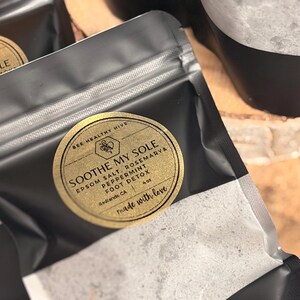 Puede incluir: Primer plano de bolsas negras con etiquetas doradas. Las etiquetas dicen "SOOTHE MY SOLE" con sal de Epsom, romero, menta y ingredientes de desintoxicaci&oacute;n de pies. Tambi&eacute;n se ve el texto "BEE HEALTHY HIVE" y "made with love". Las bolsas est&aacute;n llenas de una sustancia blanca.