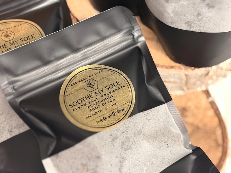 Puede incluir: Primer plano de bolsas negras con etiquetas doradas. Las etiquetas dicen "SOOTHE MY SOLE" con sal de Epsom, romero, menta y ingredientes de desintoxicaci&oacute;n de pies. Tambi&eacute;n se ve el texto "BEE HEALTHY HIVE" y "made with love". Las bolsas est&aacute;n llenas de una sustancia blanca.